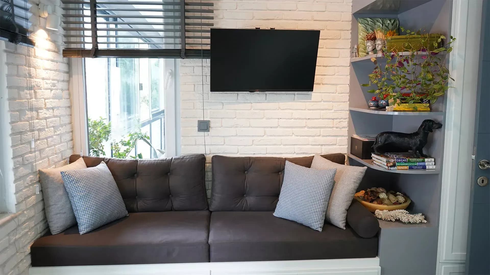 Beyaz tuğla duvarlı bekleme köşesi: kahverengi oturma bankı, yastıklar ve kitaplı raflar
