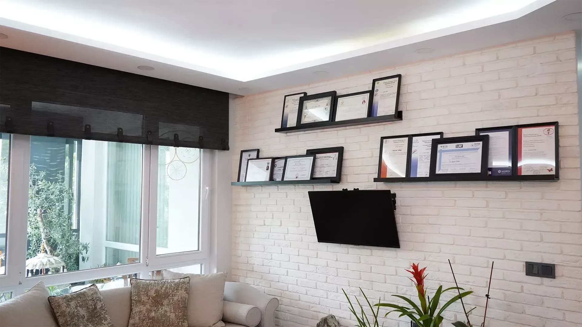 Klinik bekleme salonunda beyaz tuğla duvara asılı çerçeveli sertifikalar ve krem kanepe