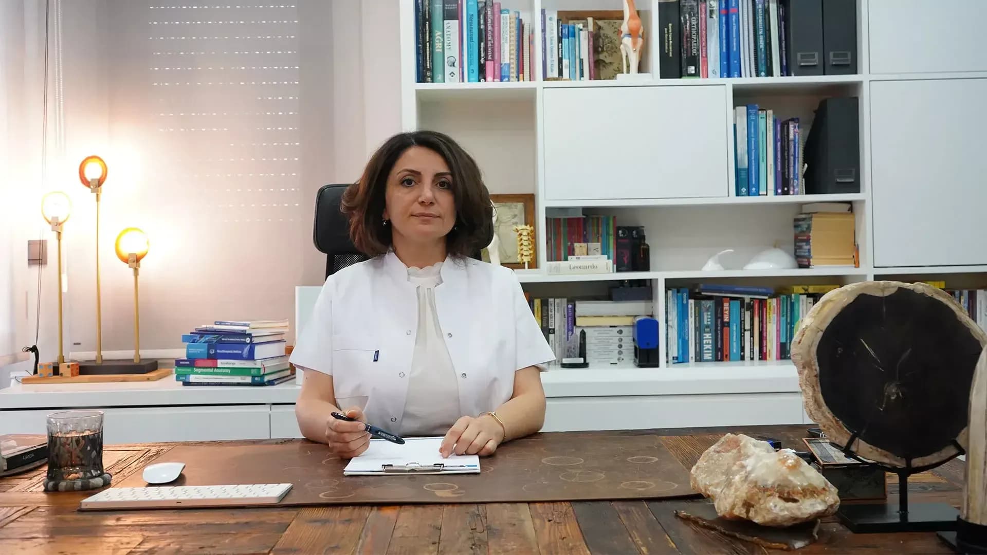 Uzm. Dr. Ayşen Türk beyaz önlüğüyle çalışma masasında; arkasında tıp kitapları ve anatomi modelleri