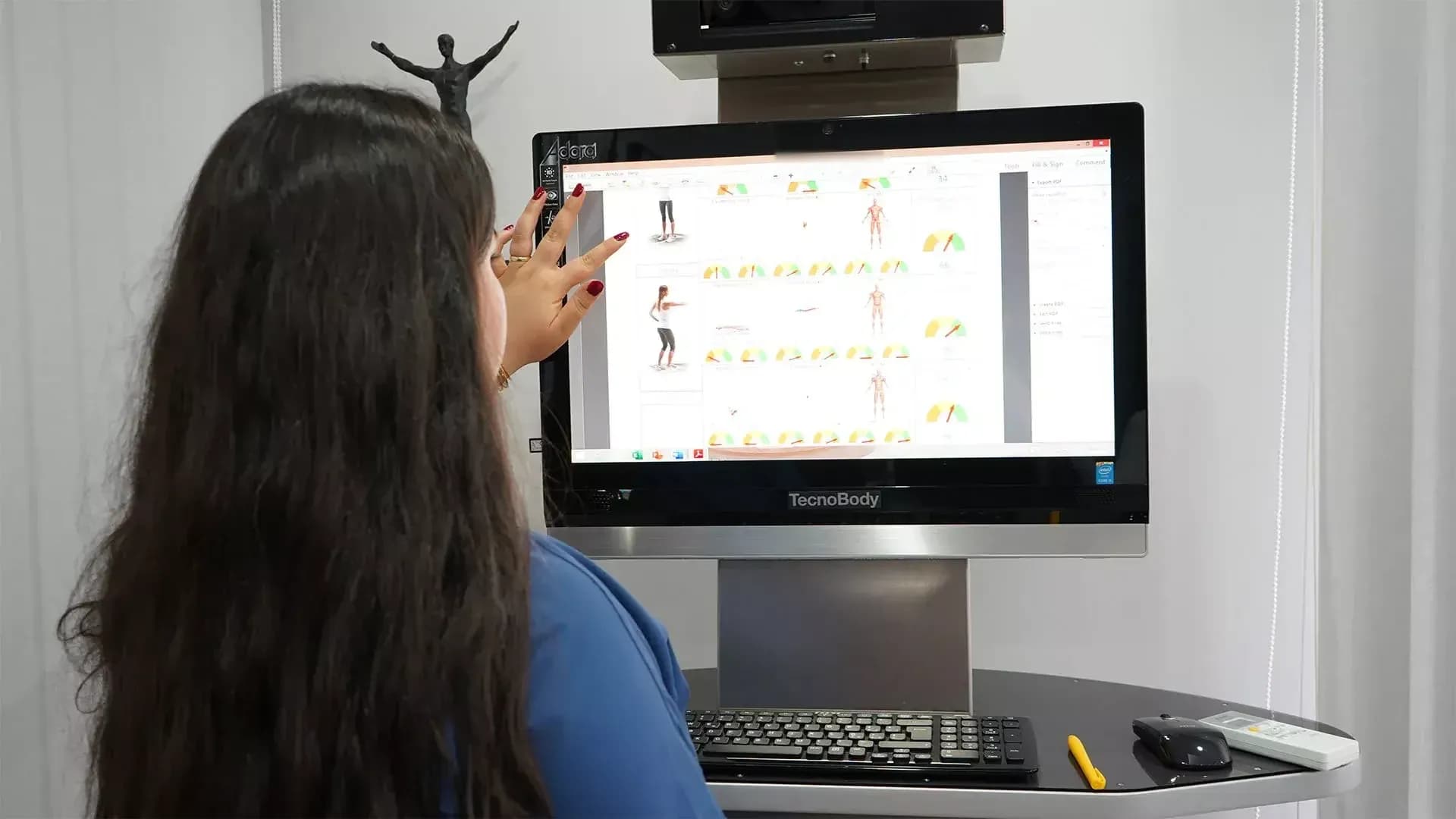 Mavi üstlü bir kişi, duruş analizi raporunun açık olduğu masaüstü bilgisayar ekranını eliyle işaret ediyor