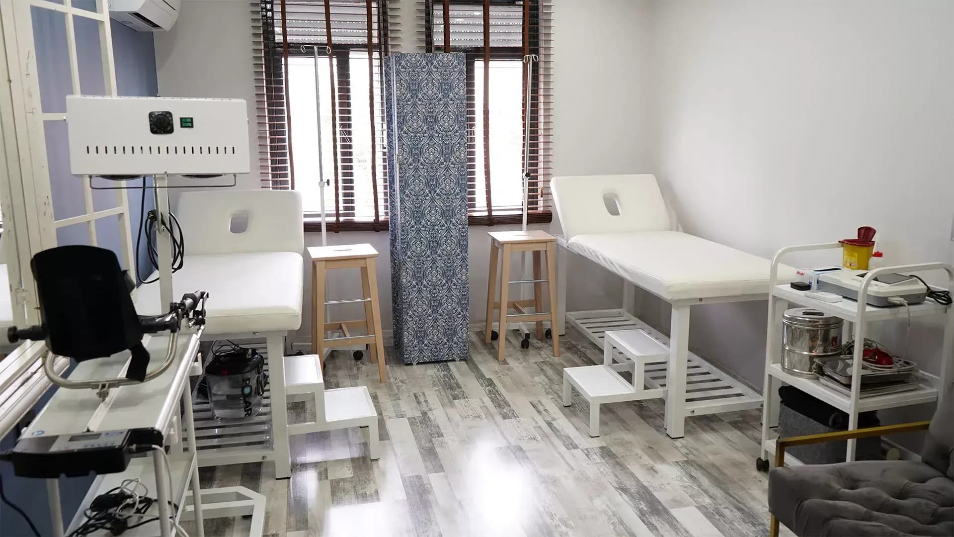 İki muayene sedyesi, ayaklı stant üzerinde ışık-ısı cihazı, serum askıları ve önde koşu bandı bulunan uygulama odası